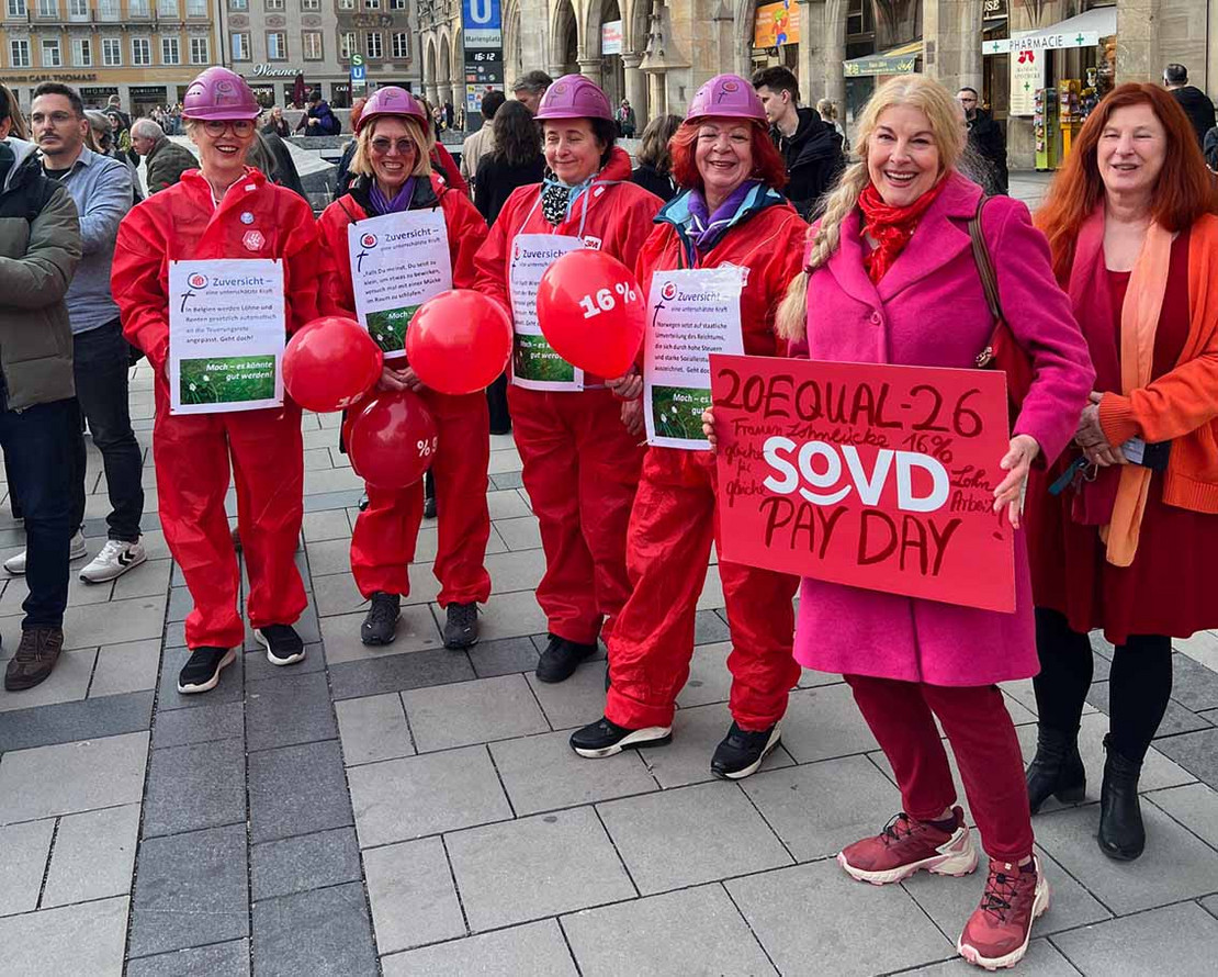 Gruppe mit rot gekleideten Personen, eine Frau präsentiert ein Plakat mit der Aufschrift "Equal Pay Day"