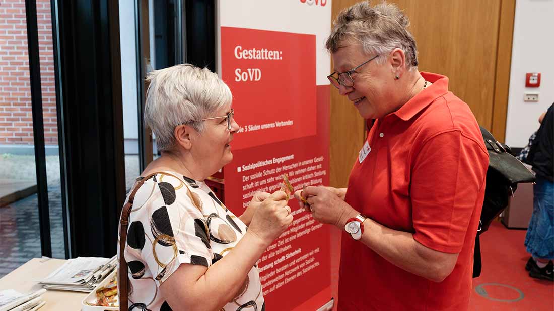 Zwei Personen unterhalten sich. Im Hintergrund ein SoVD-Aufsteller. 